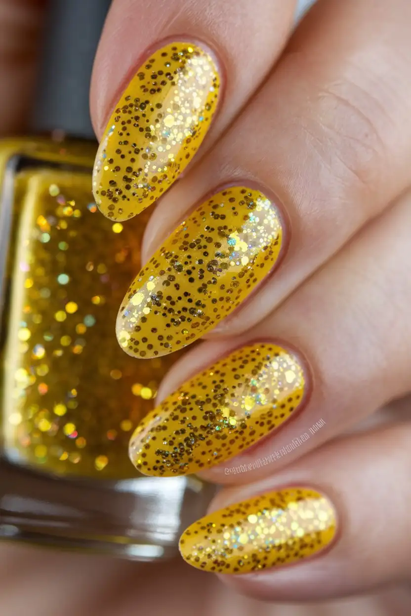 A close-up shot of a hand with almond-shaped nails painted in a bright, sunny yellow nail polish with a layer of fine gold glitter sprinkled on top. The glitter catches the light, creating a dazzling effect. The finish is glossy and the lighting is bright, highlighting the sparkle. The background is a blurred image of golden glitter falling