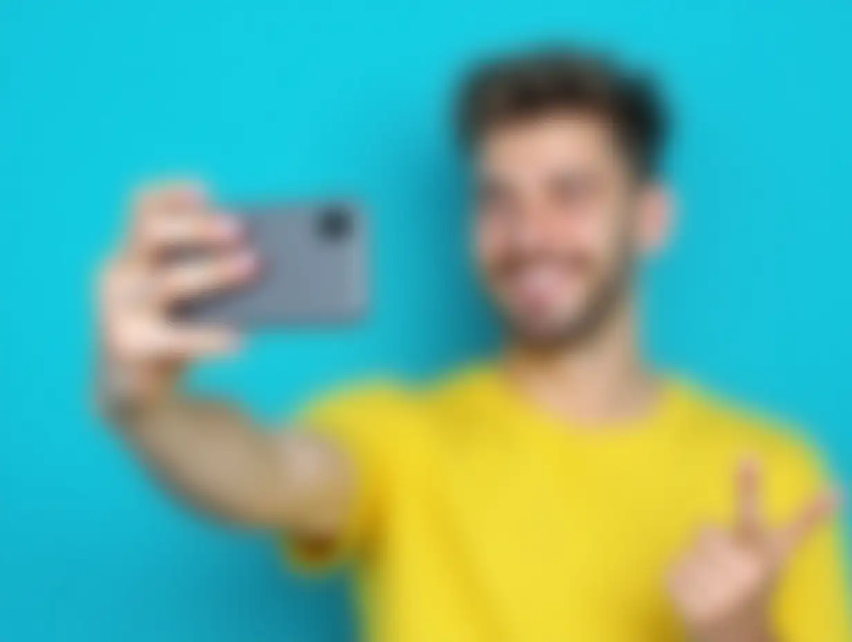 Happy young man taking a selfie with a smartphone against a blue background, wearing a yellow t shirt, smiling and making a peace sign, showcasing joy and carefree attitude